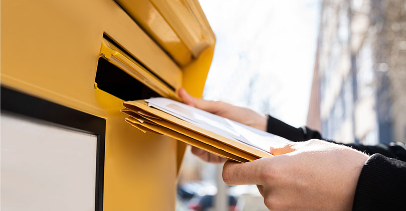 Ein Mensch wirft einen schmalen Papp-Versandumschlag in einen gelben Briefkasten